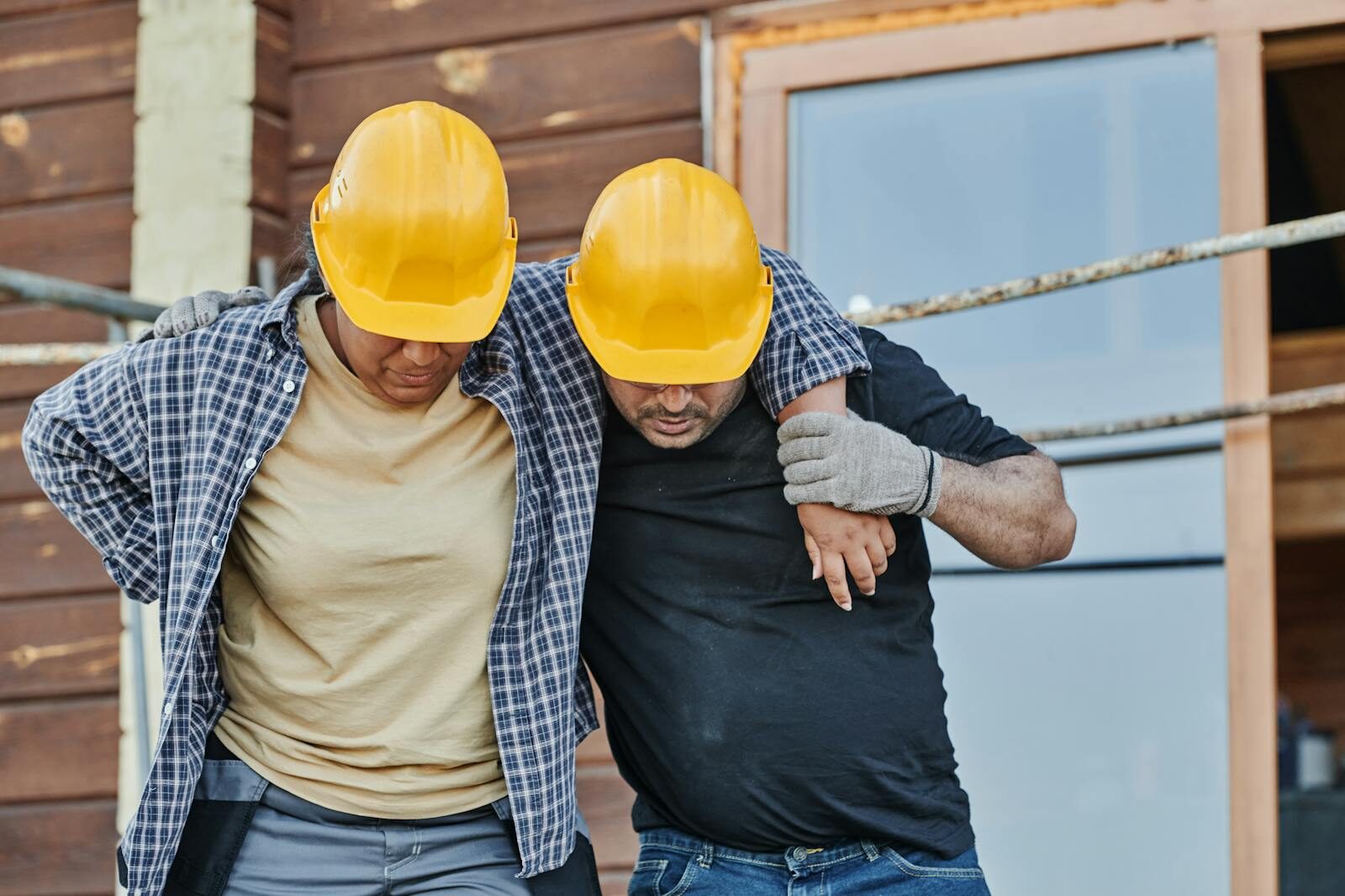 Two construction workers in hard hats assisting each other at a building site, highlighting teamwork.